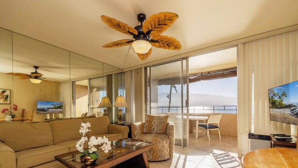 A spacious living room with a large sofa, a coffee table with flowers, a patterned armchair, and a ceiling fan.