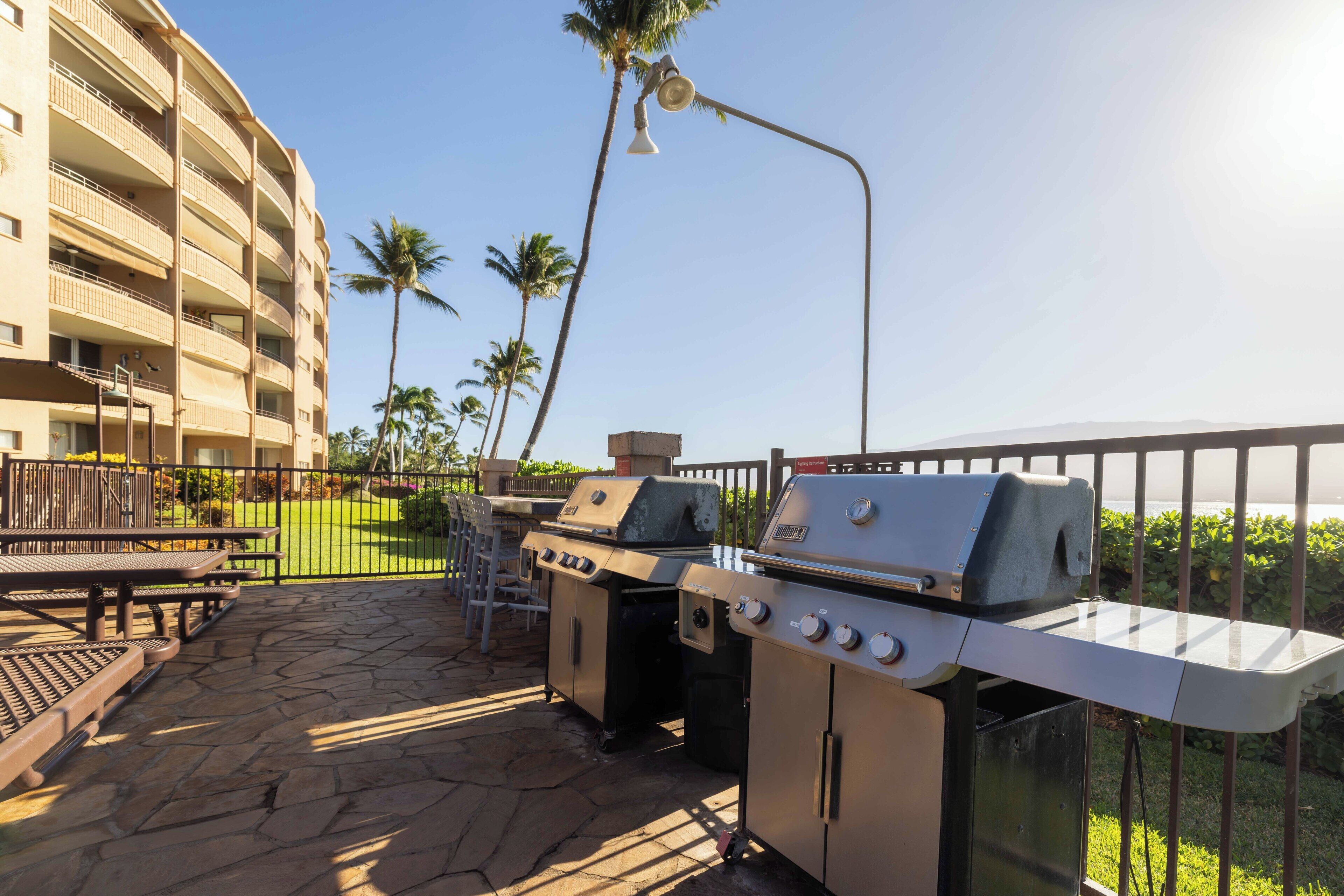 An outdoor patio with stone flooring, two barbecue grills, a wooden bench, and a view of a multi-story building with balconies.
