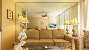 A living room with a beige sofa, a glass coffee table, and two lamps. There is a ceiling fan, a television, and a bookshelf.