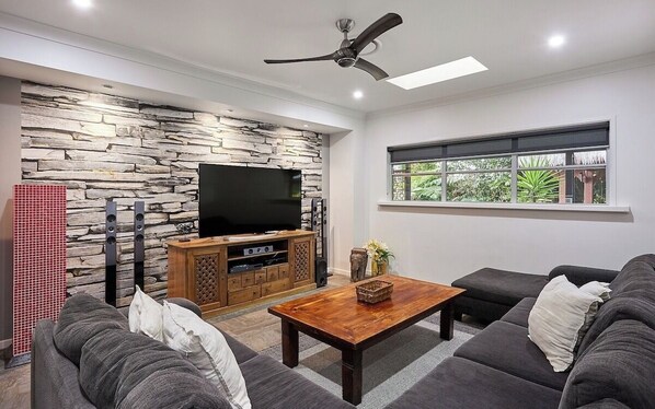 A modern living room with a stone wall, a flat-screen TV, a wooden entertainment unit, a ceiling fan, and a large window with a view of greenery.