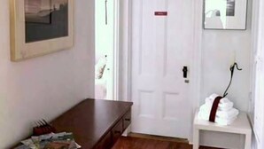 Een kamer met een houten dressoir, een ingelijst schilderij aan de muur, een witte deur en een klein wit bankje met een rode lint.