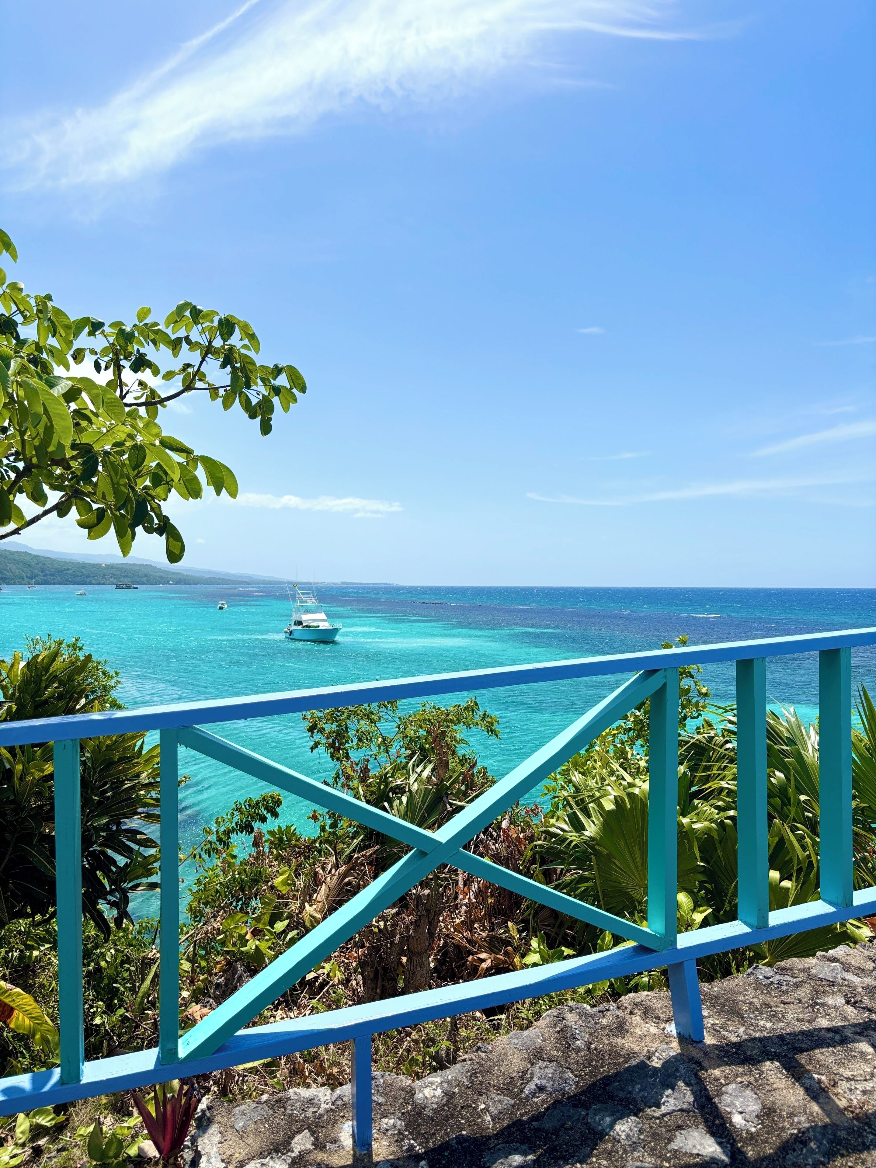 Une balustrade turquoise surplombe une mer d’un bleu limpide, avec un bateau visible au loin.