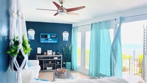 A room with a ceiling fan, a TV mounted on the wall, a sofa, a coffee table, and a view of the ocean through large windows. - Oceanview 2BR Condo Walk to Mahogany Beach Ocho Rios (Ocho Rios)