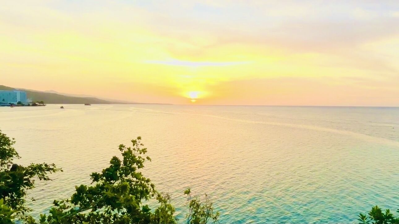 Une vue sur la côte, avec un coucher de soleil sur l’eau, un bâtiment sur la gauche et de la végétation au premier plan.