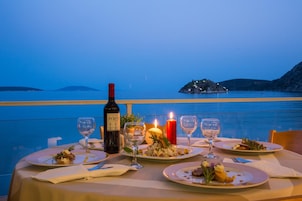 Una mesa servida para cenar con platos de comida, copas de vino y una vela, con vistas a una masa de agua y una isla distante.
