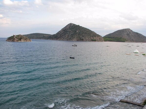 A beach with calm waters, a small island with a building, and distant hills. - Flisvos Hotel Tolo (Nafplio)
