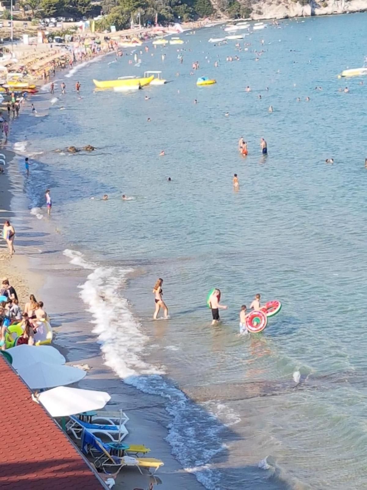A crowded beach with people swimming and walking along the shore, boats, and umbrellas.