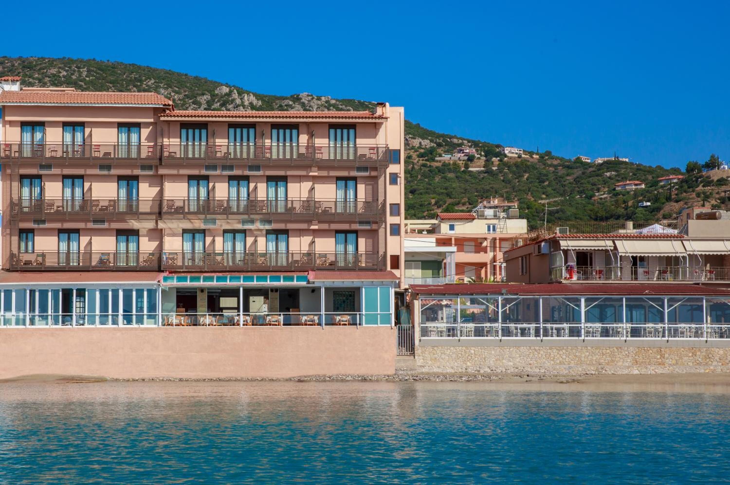 A multi-story hotel with balconies, a pool area, and a view of the sea and hills in the background.