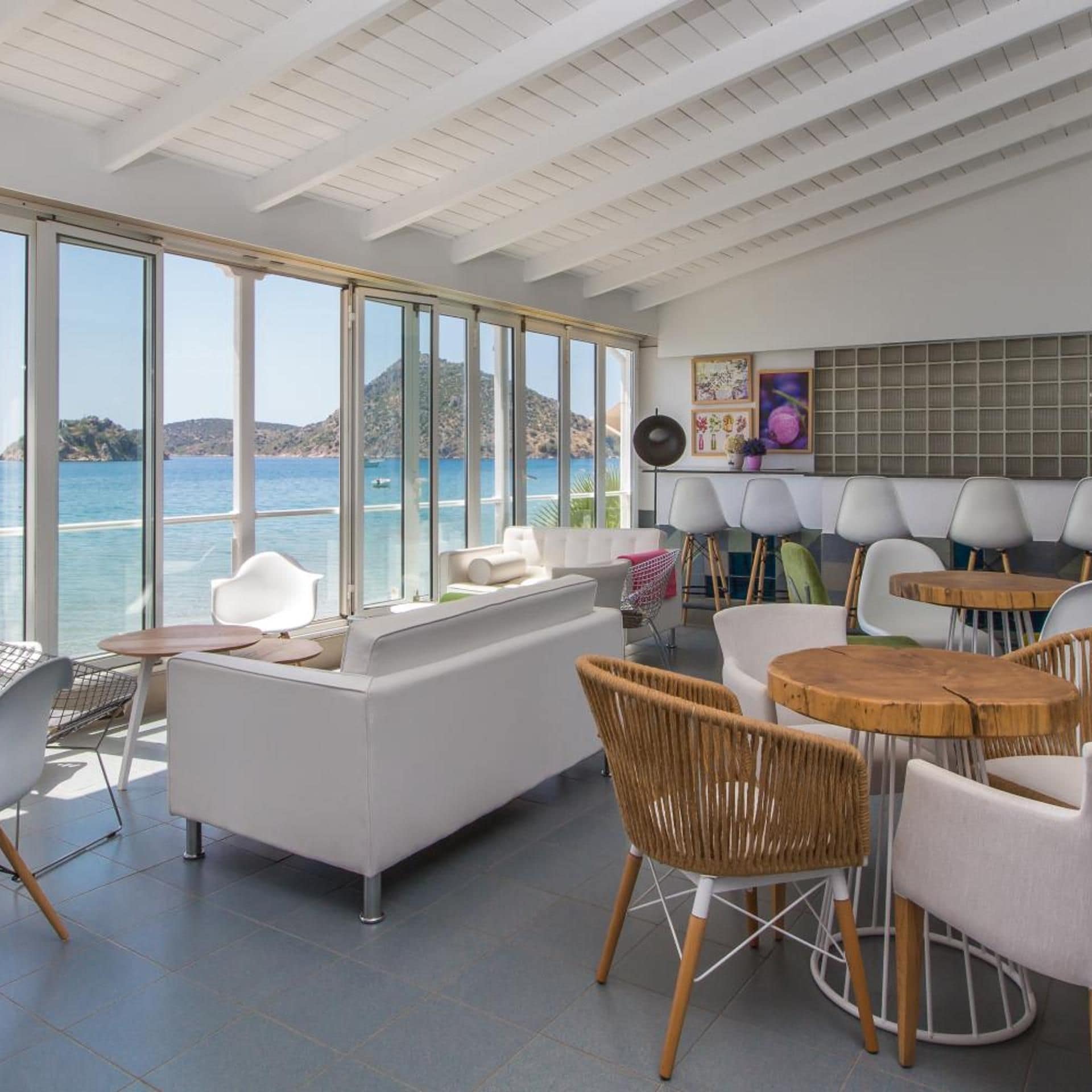 A modern interior with a large window overlooking a body of water, a seating area with a sofa and chairs, and a dining setup with tables and chairs.
