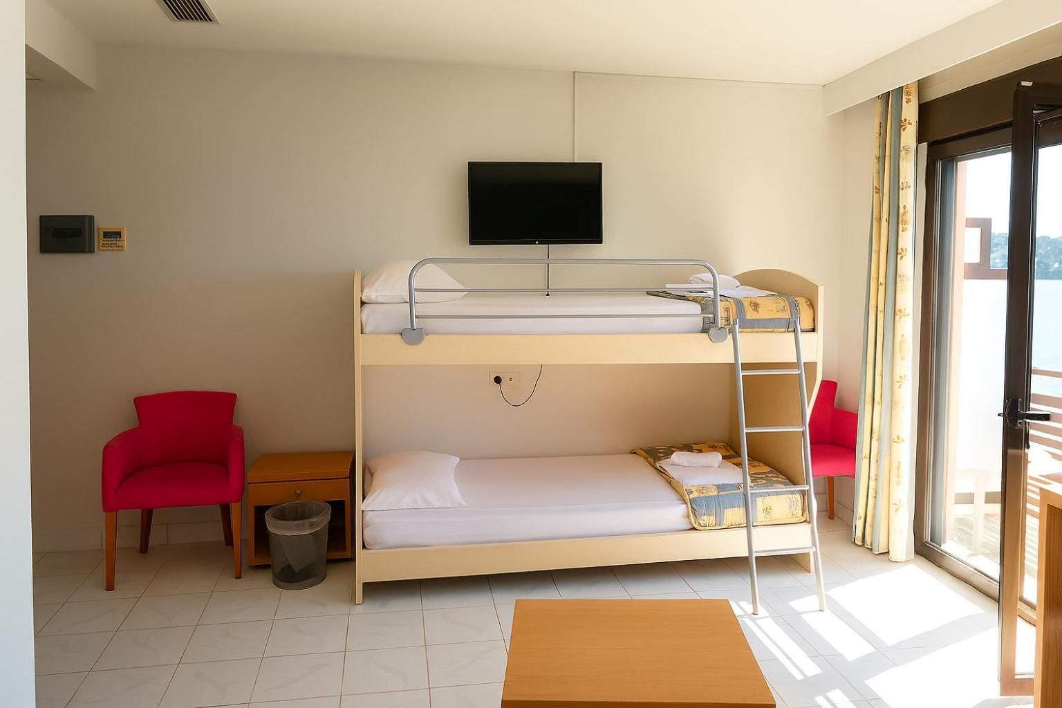 A bunk bed room with a television above it, a small table, two red chairs, and a window with curtains.