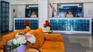 A hotel lobby with a reception desk, a sofa with floral arrangements, and a large window with blue-tiled walls. - Flisvos Hotel Tolo (Nafplio)