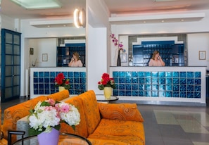 A hotel lobby with a reception desk, a sofa with floral arrangements, and a large window with blue-tiled walls. - Flisvos Hotel Tolo (Nafplio)