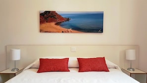 A hotel room with a bed, two red pillows, white bedding, and a painting of a beach on the wall.