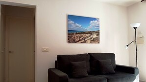 Ein Wohnzimmer mit einem dunkelgrauen Sofa, einem Wandbild von einer Strandlandschaft, einer Tür und einer Stehlampe.