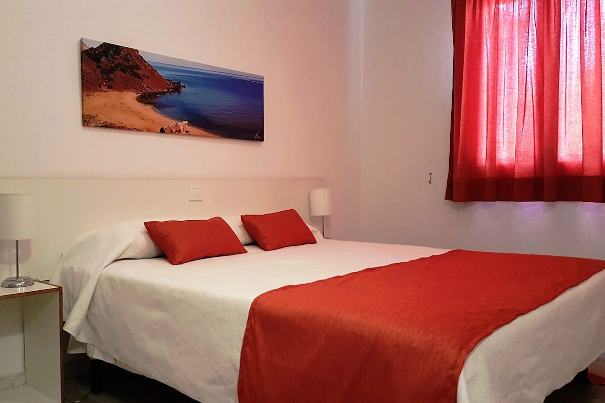 A hotel room with a bed, two bedside lamps, a red blanket, a red curtain, and a painting of a beach on the wall.