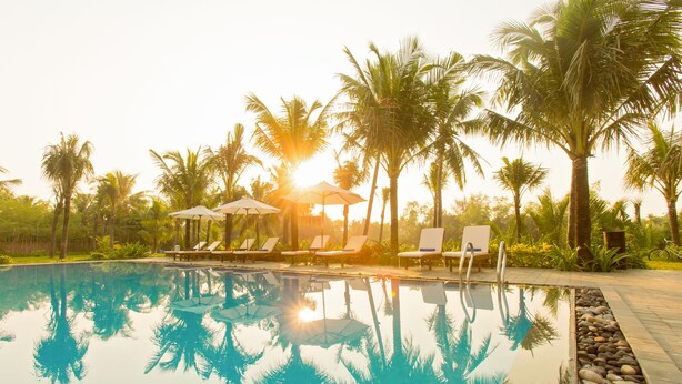 A pool with clear water reflecting palm trees and a sun setting in the background. - Hoi An Waterway Resort (Hoi An)