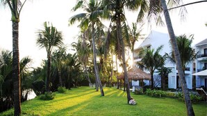 A tropical resort with palm trees, a thatched-roof structure, and a white building with balconies. - Hoi An Waterway Resort (Hoi An)