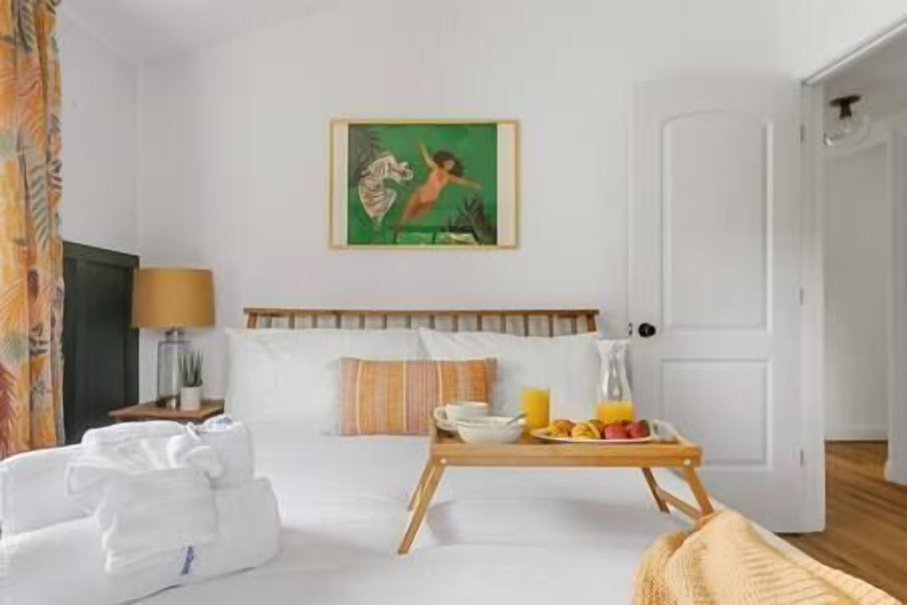 Habitación de hotel con cama, mesita de noche con lámpara, un cuadro en la pared y una bandeja de desayuno con zumo de naranja y croissants.