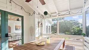 Une table en bois avec une assiette de nourriture et un verre, un plafond blanc avec des luminaires suspendus, et une vue sur les arbres à travers de grandes fenêtres.