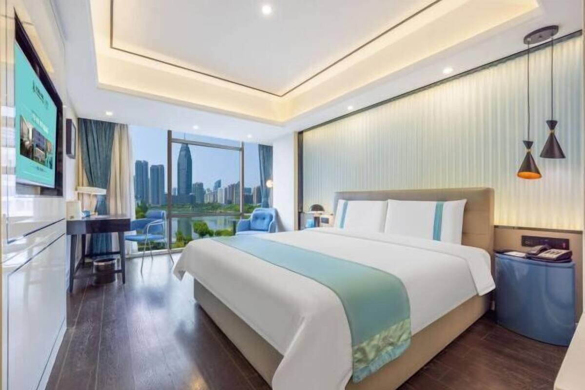 Une chambre d’hôtel moderne, dotée d’un grand lit, d’un bureau, d’une chaise, d’une petite table et offrant une vue sur le paysage urbain.