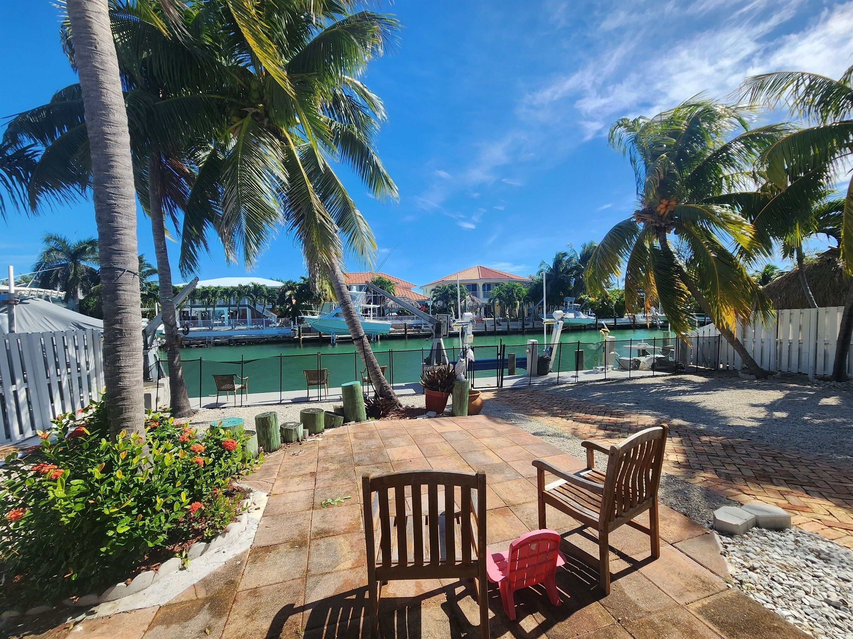 Un patio con sillas y una mesa de madera, rodeado de palmeras y vistas a un puerto deportivo con barcos.