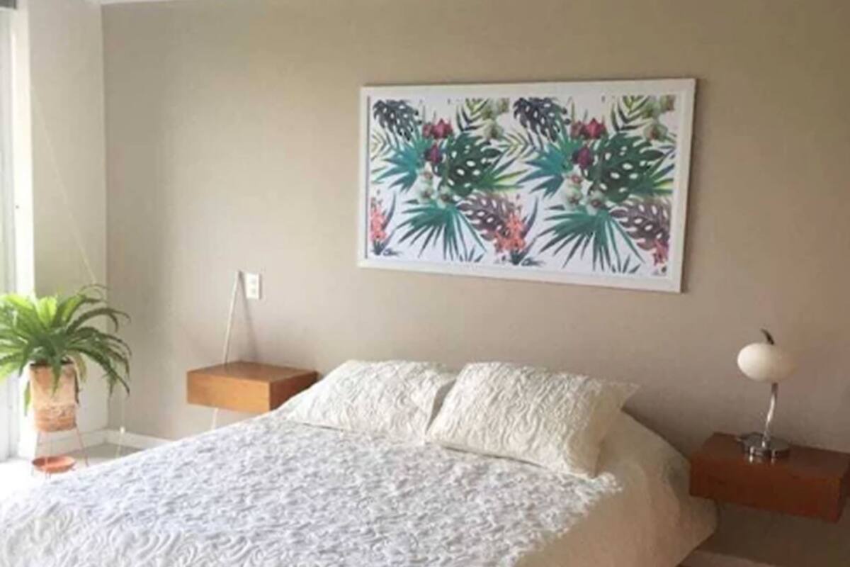 A bedroom with a large bed, a nightstand, a wall-mounted lamp, and a potted plant.