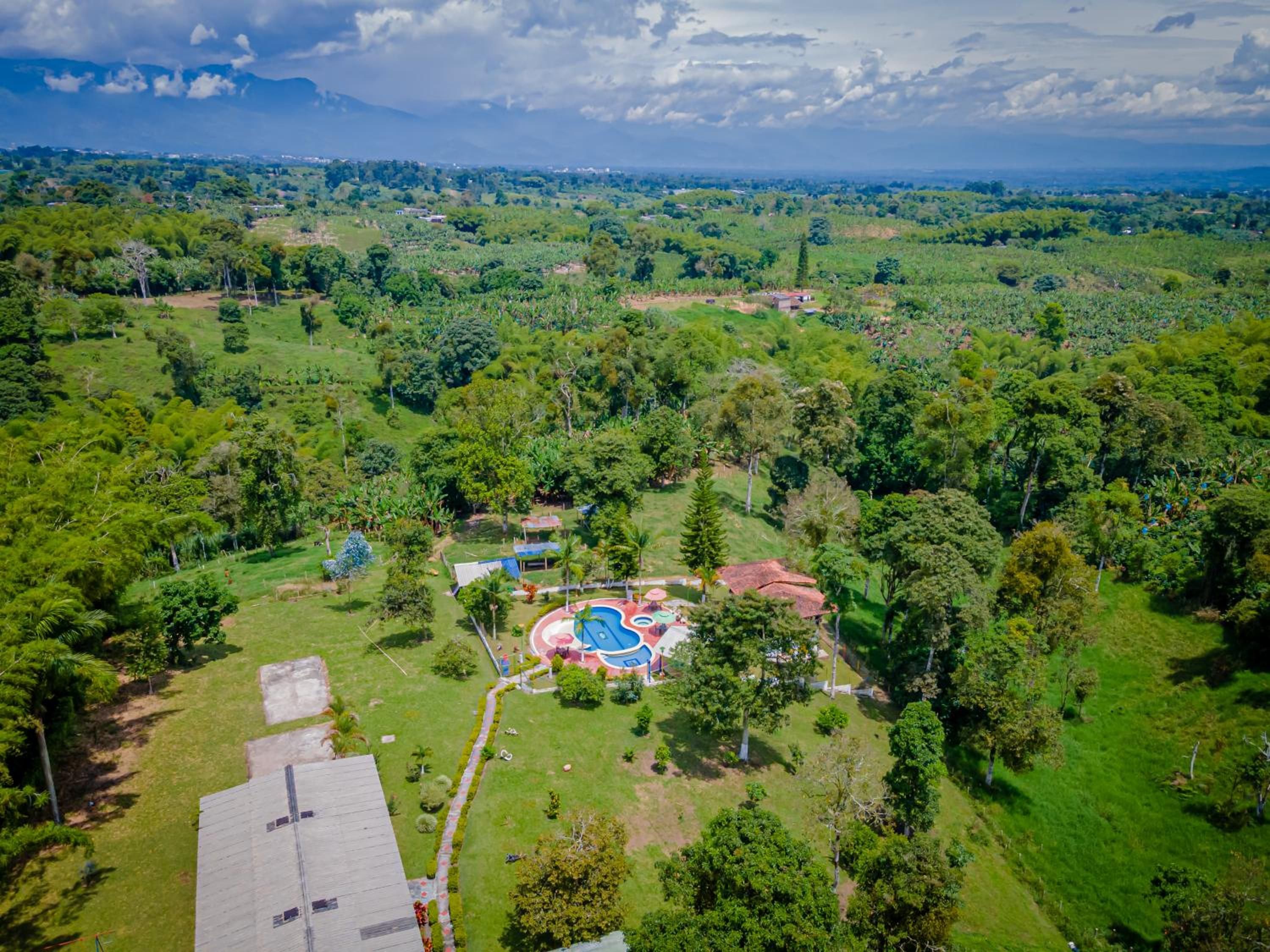 Vista aérea de un complejo turístico con piscina, rodeado de exuberante vegetación y colinas al fondo.