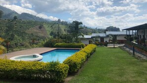 A pool surrounded by well-maintained grass and bushes, with a mountain backdrop and a building with a sloped roof.