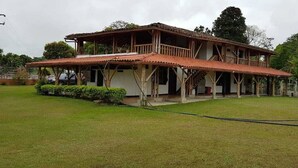 A two-story wooden house with a balcony, surrounded by a grassy lawn and trees.