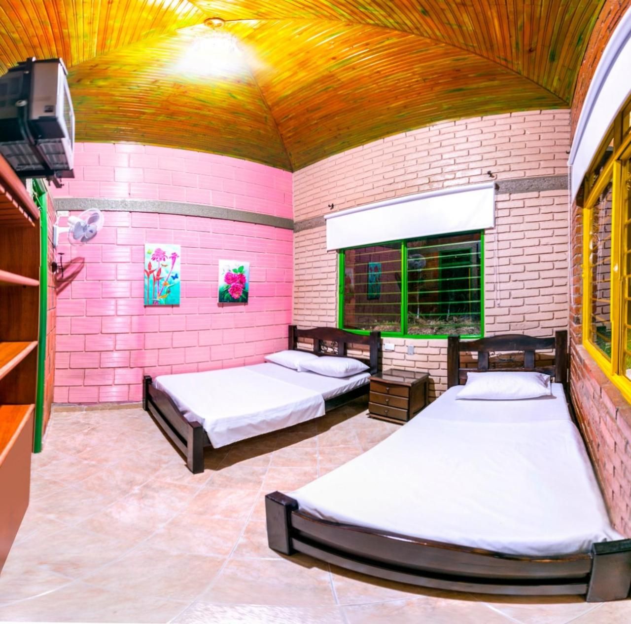 A room with two beds, a window with green bars, a fan, and a pink brick wall.