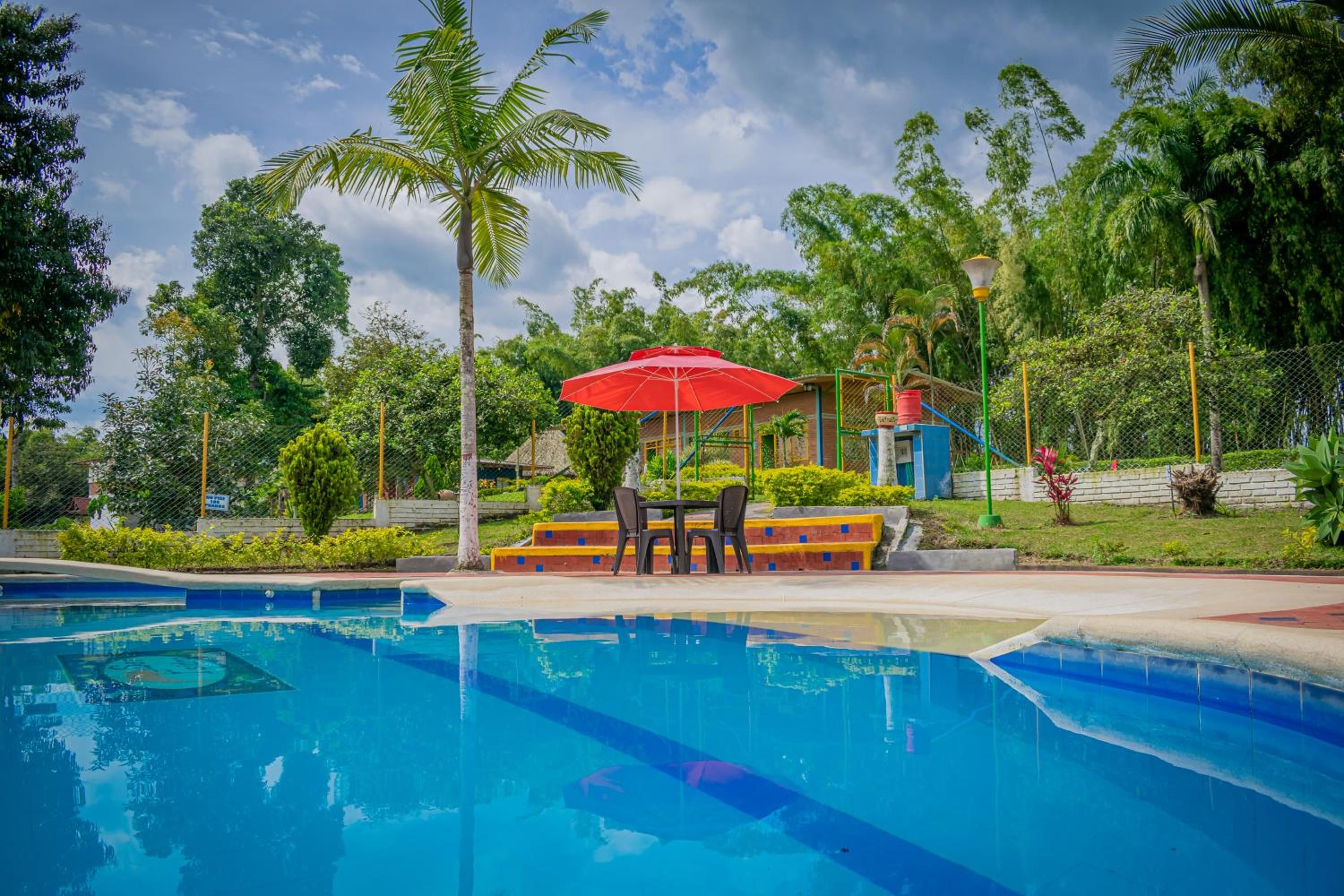 A swimming pool with a red umbrella and two chairs, surrounded by greenery and a clear sky.