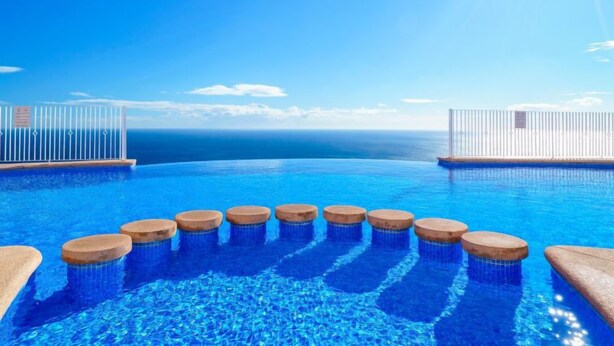 Ein Pool ohne Ende mit Holzstufen, die ins Wasser führen, umgeben von weißen Geländern und einem ungehinderten Blick auf das Meer unter einem strahlenden Himmel.