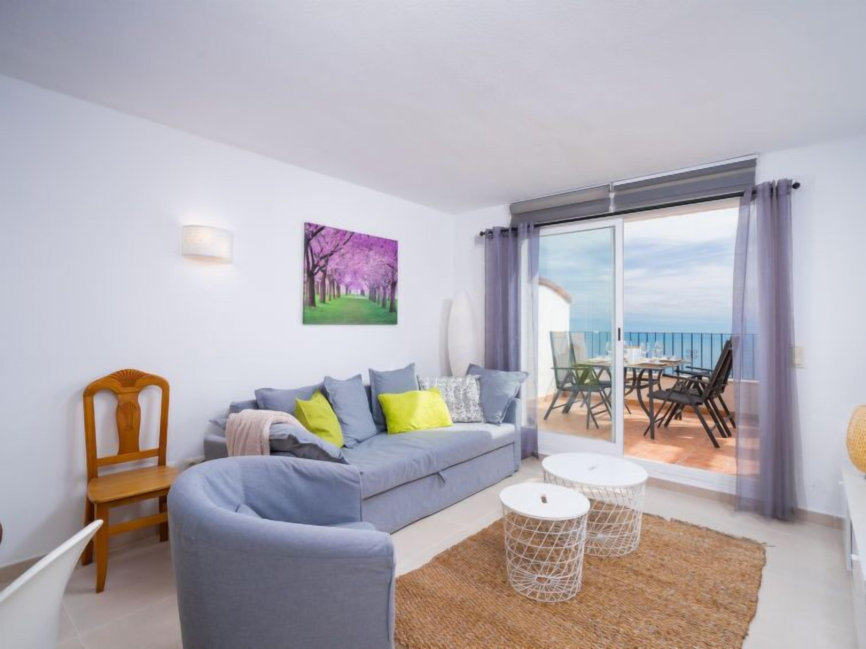 Ein modernes Wohnzimmer mit einem grauen Sofa, einem Holzstuhl und zwei weißen runden Tischen. Ein großes Fenster bietet Blick auf das Meer.