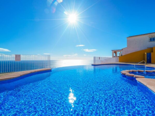 A clear blue infinity pool with a view of the ocean, a sunlit sky, and a building with a yellow facade. - Pueblo Panorama by Interhome (Moraira)
