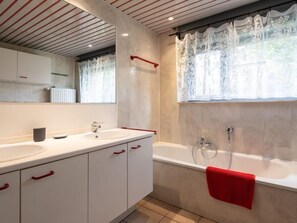 A modern bathroom with two sinks, a large mirror, a bathtub, and a window with lace curtains.