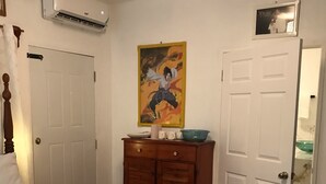 A bedroom with a bed, a wooden dresser, a painting, and a door leading to a bathroom. - Centrally located room, in a house with free Wifi. (Port Antonio)