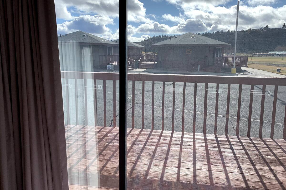 A view from a balcony with a wooden railing, overlooking a parking area with two buildings and a clear sky with clouds.