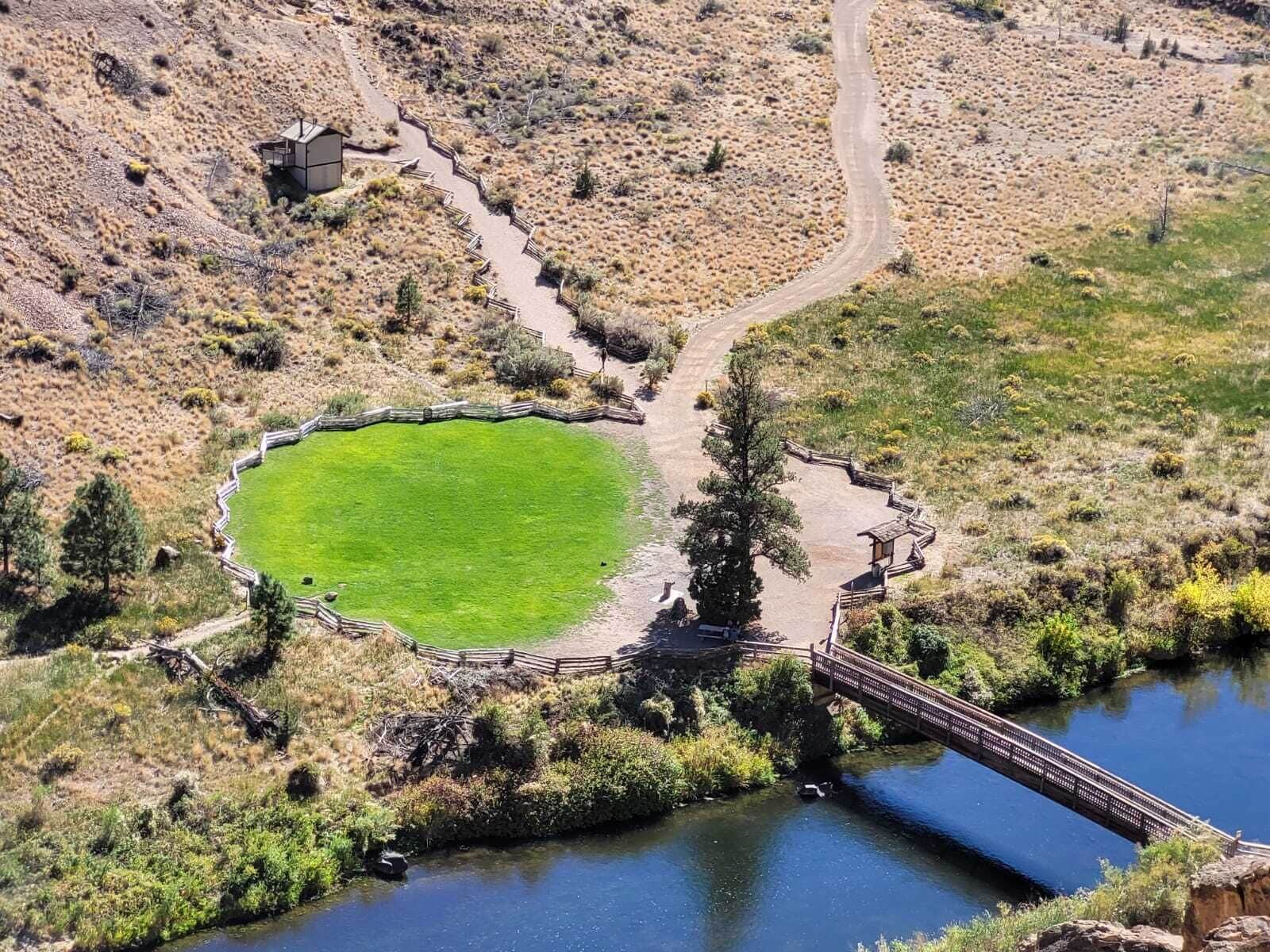 An aerial view of a park with a circular green area, a bridge over a water body, and a dirt path leading to a structure.