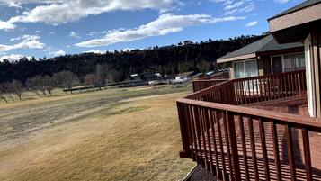 Un balcón de madera con vistas a un campo cubierto de hierba, donde se distinguen casas y árboles a lo lejos.