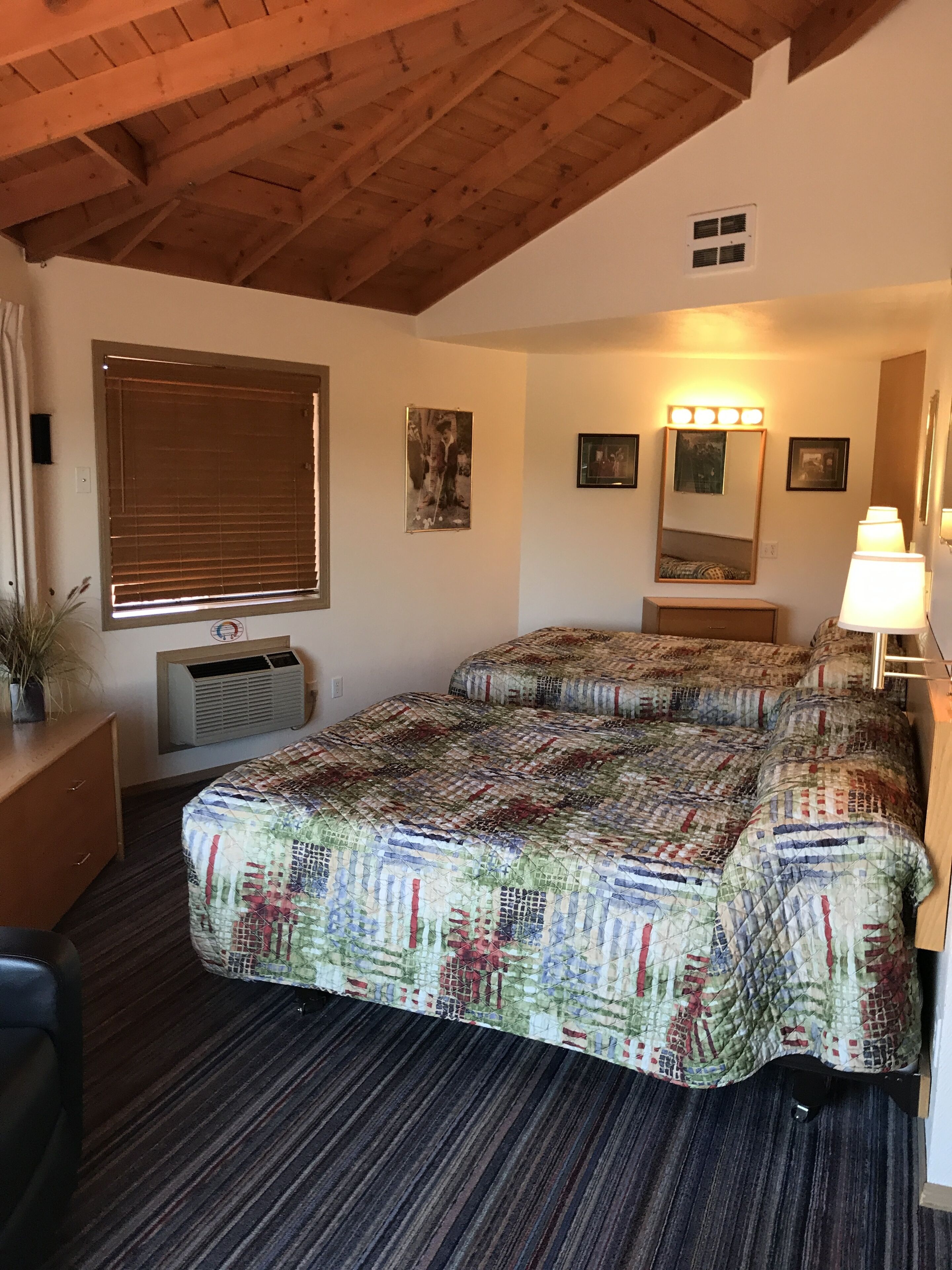 Quarto com teto de madeira, uma cama grande com colcha colorida, mesa de cabeceira com abajur e janela com persianas.