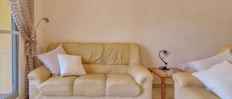 A beige leather sofa with cushions, a small side table with a lamp, and a window with curtains.