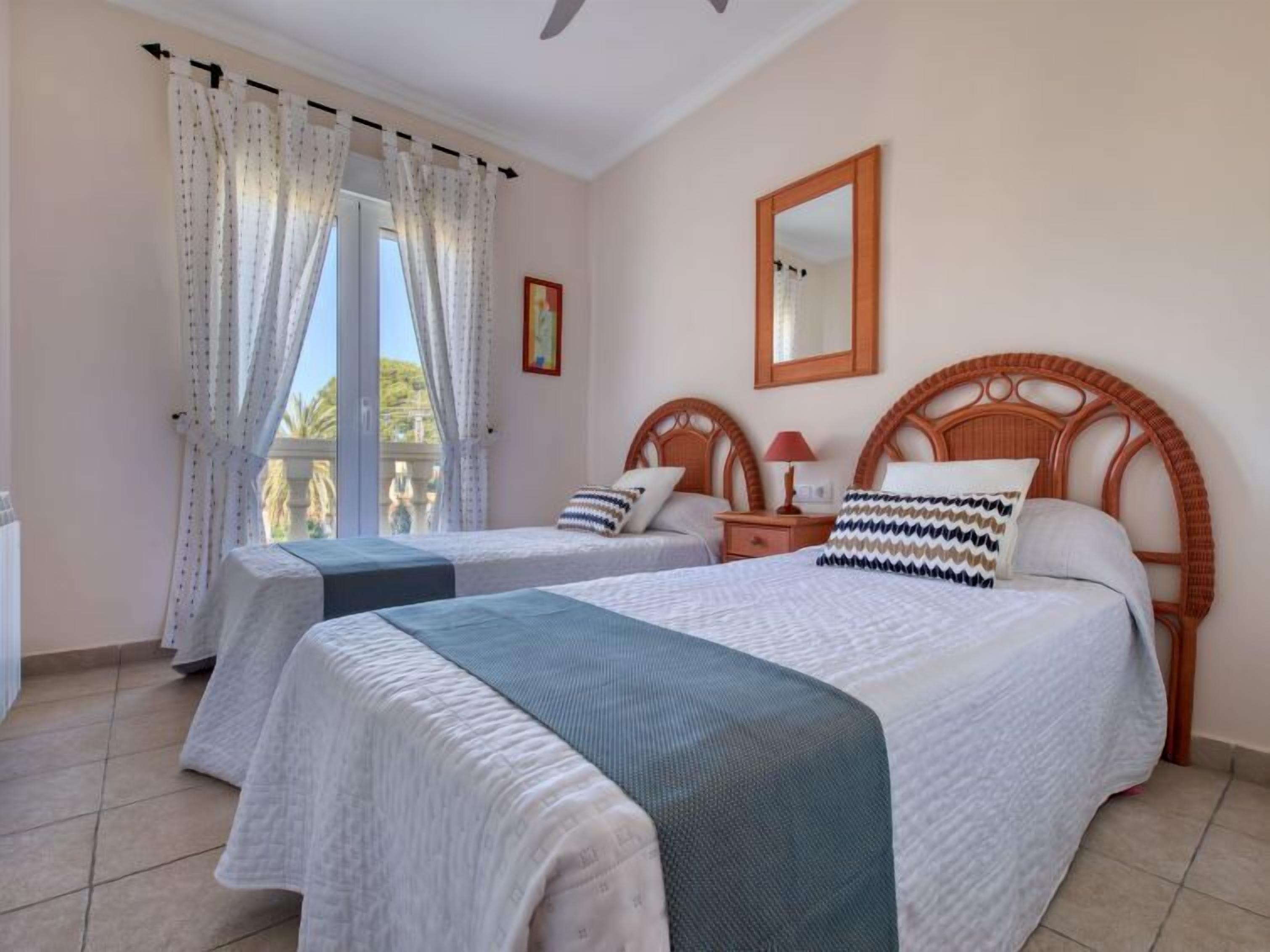 A hotel room with two beds, a balcony, a ceiling fan, a mirror, and a window with curtains.