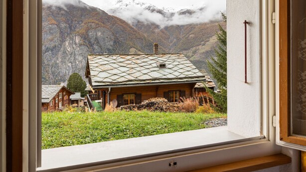 A rustic cabin with a stone roof, surrounded by lush greenery and mountains in the background.