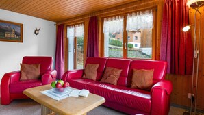 A cozy room with a red leather sofa, a wooden table with a bowl of fruit, and large windows with curtains.