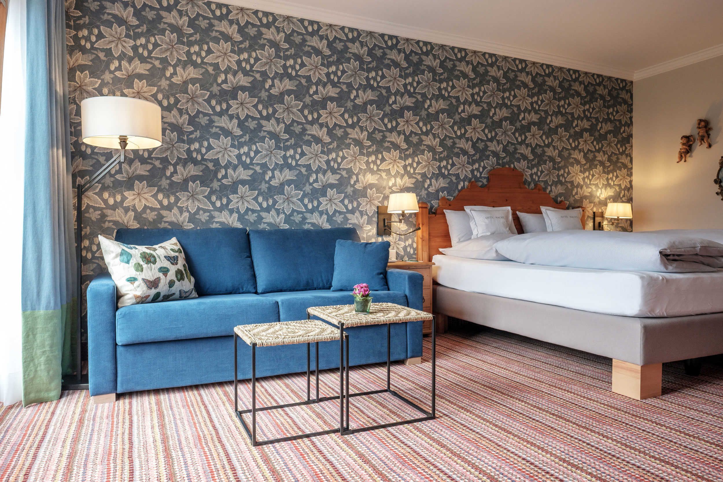 Habitació d'hotel amb un sofà blau, un llit, dues taules petites, una catifes amb dibuixos i un paper pintat floral.