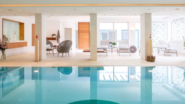 A modern indoor pool area with a clear view of the pool, lounge chairs, and a seating area with a view of the outside.