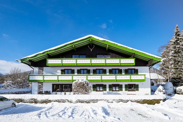 A two-story building with a green roof and white walls, surrounded by snow. - Suite de Luxe - Landhaus am Stein (Bad Wiessee)