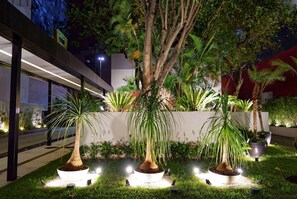Un espace extérieur bien éclairé, avec un bâtiment moderne, un grand arbre et plusieurs plantes en pot.