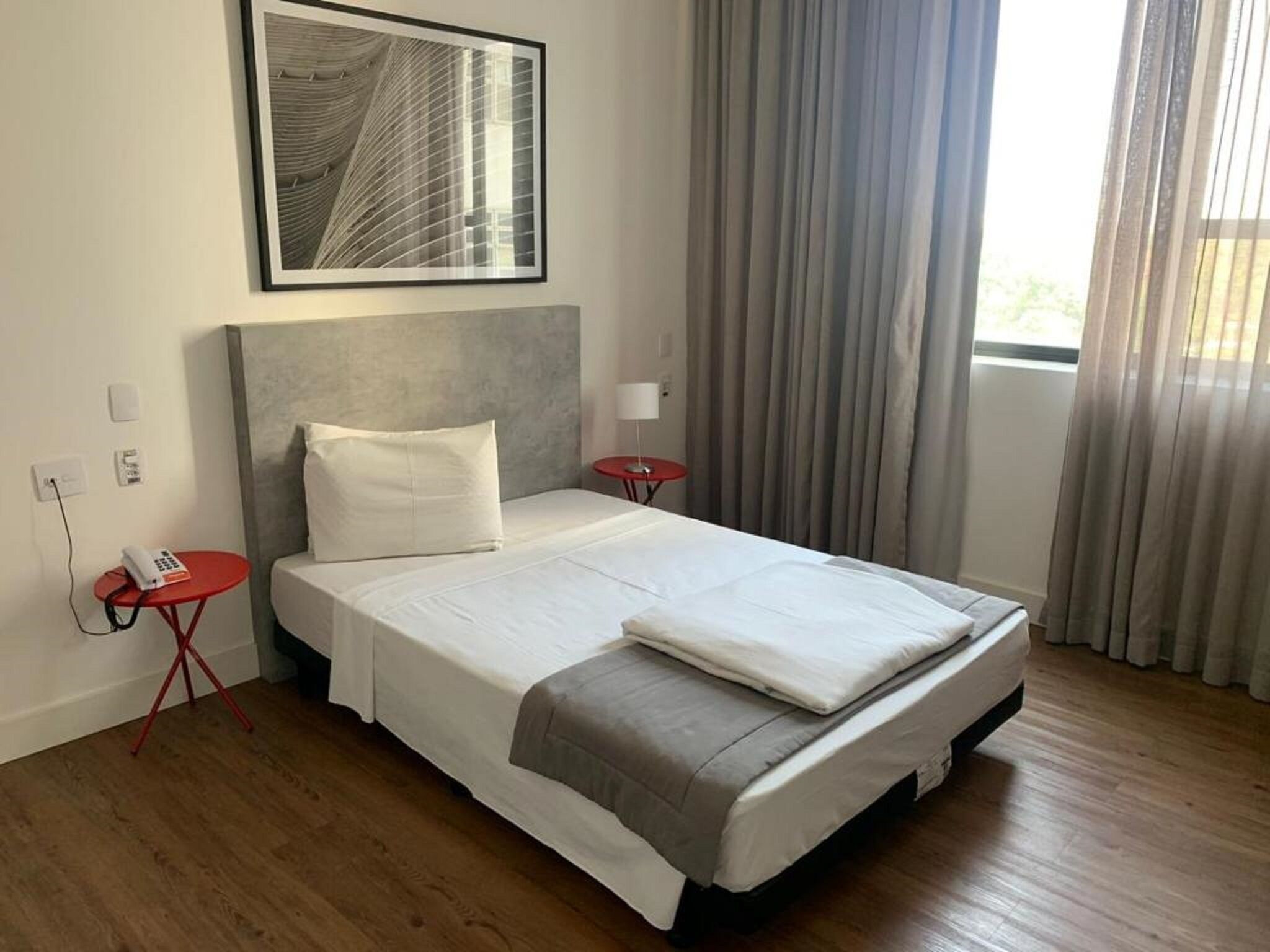 Une chambre d’hôtel avec un grand lit, une petite table de chevet avec un téléphone, une table rouge et une fenêtre avec des rideaux.