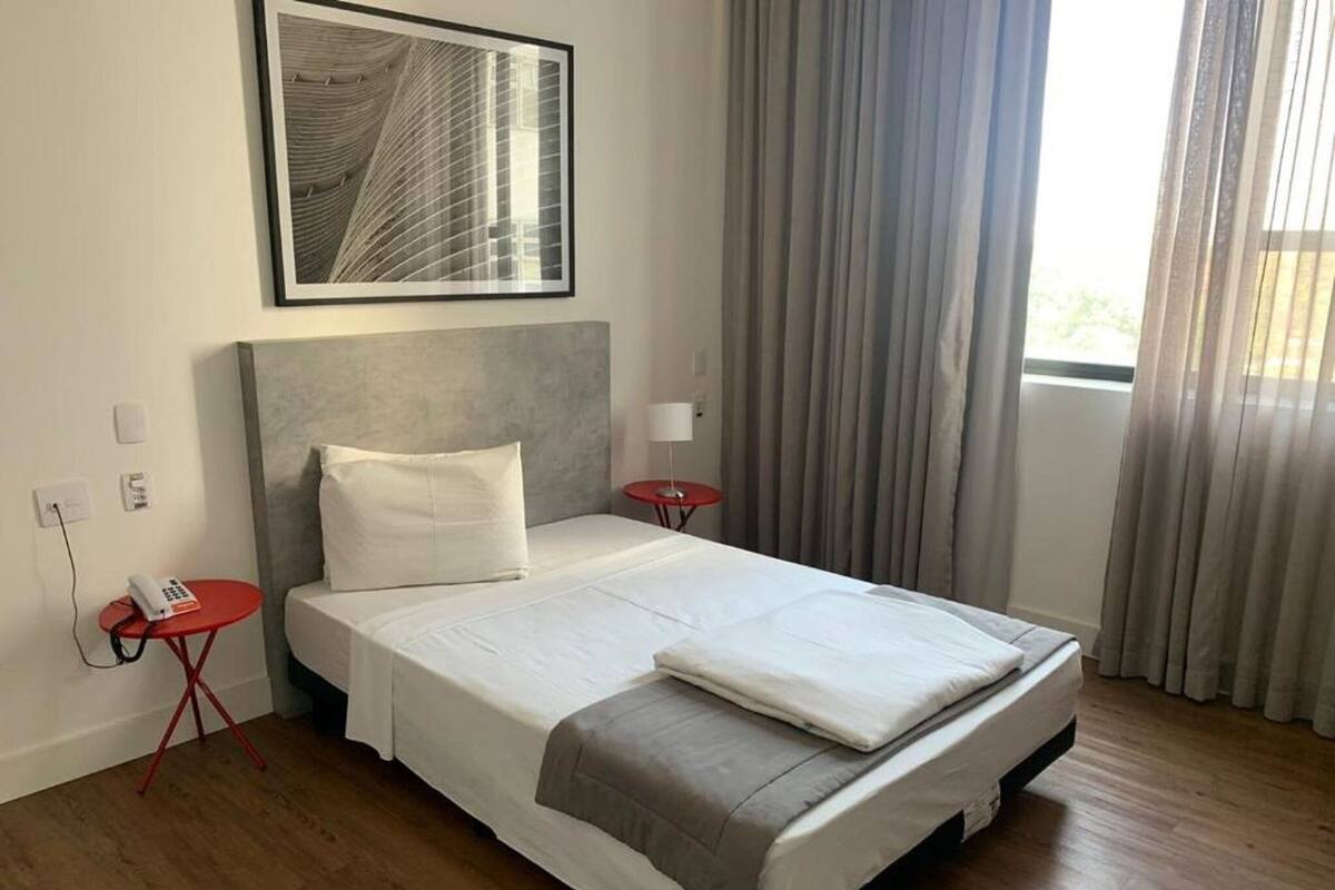 A hotel room with a large bed, a small bedside table with a phone, a red table, and a window with curtains.